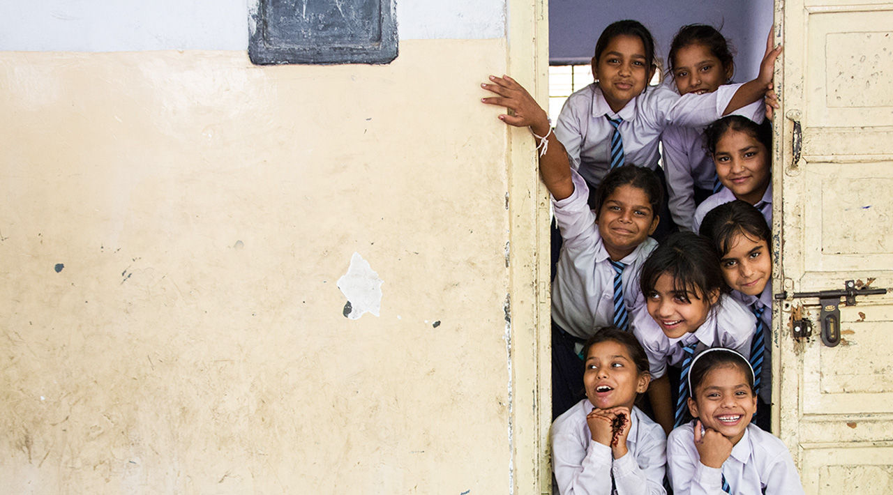 Smiling and laughing, uniformed Indian schoolchildren look out the door of a rural school. 